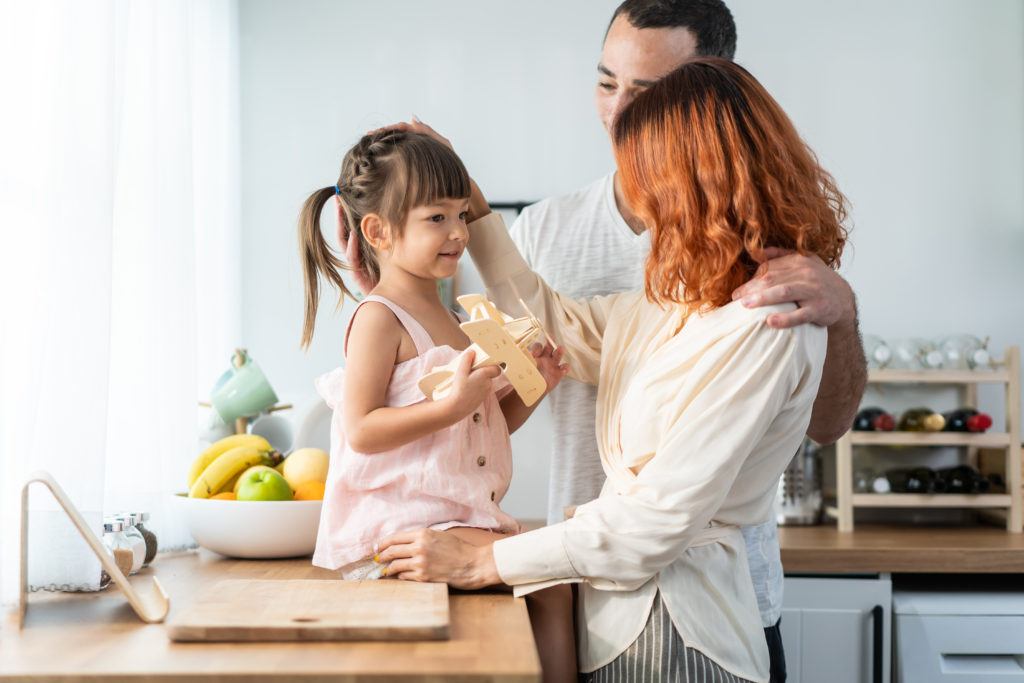 Asian attractive parents playing airplane toy with baby kid in kitchen. Happy family, young couple mother and father spend time on holiday together with daughter, prepare to cook foods for breakfast.