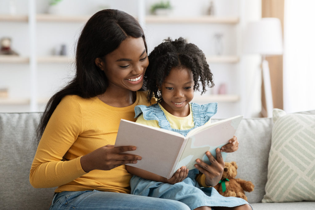 Loving black mom reading book to her cute little daughter at home, happy young african american mother and preteen female child relaxing on couch in living room, enjoying spending time together