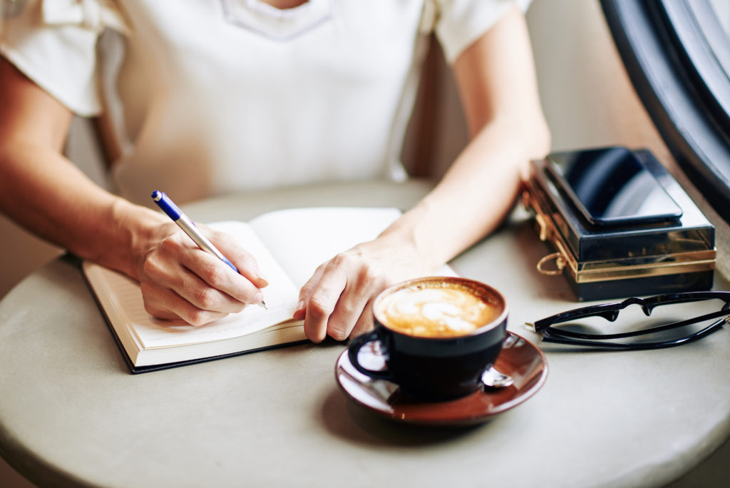 Close-up image of woman drinking cup of cappuccino and writing thoughts and ideas in gratitude journal
