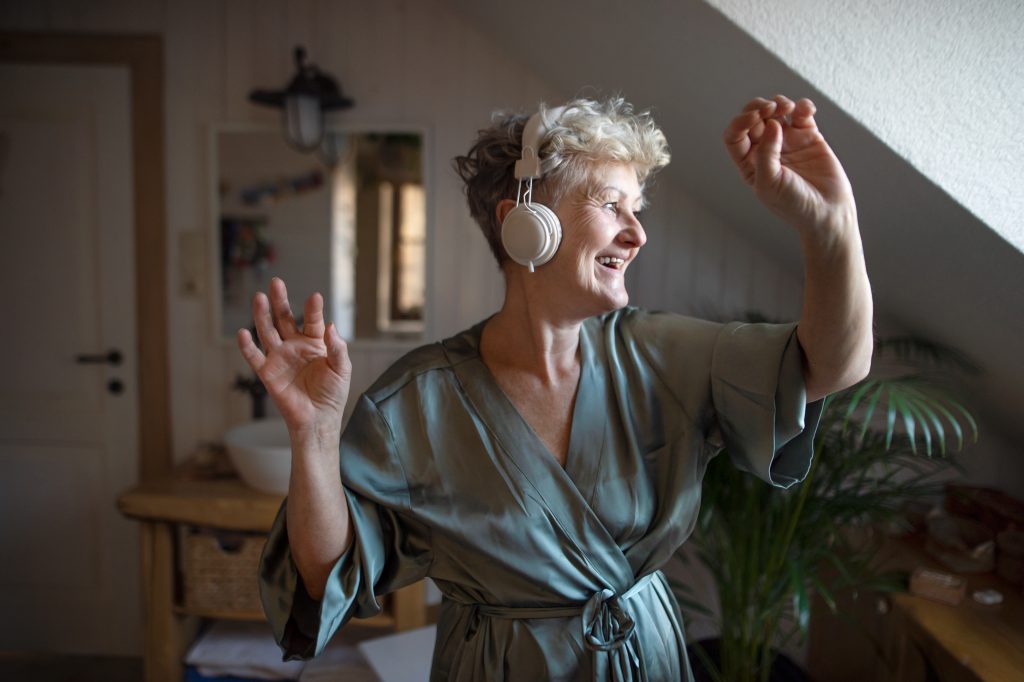 A happy senior woman with headphones dancing at home, relax and self-care concept.